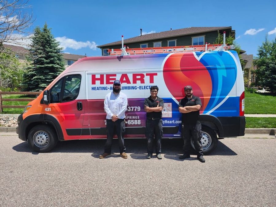 Three technicians in work attire pose in front of a colorful Heart Heating van parked on a residential street.