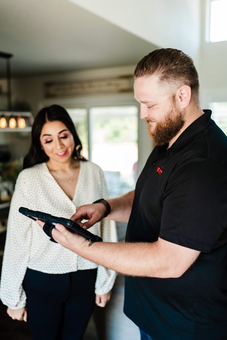 Man demonstrates a tablet device to a smiling woman in a bright home setting.