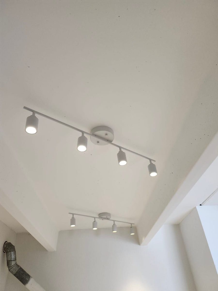 Modern white track lighting on a sloped ceiling with two bars and four cylindrical spotlights each, illuminating a bright interior, with a duct visible in the corner.