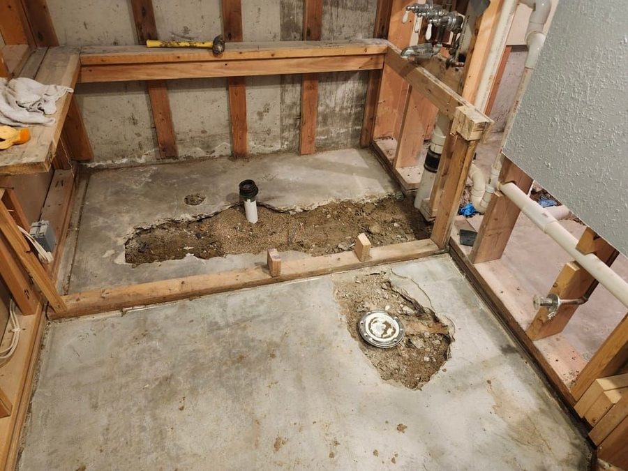 Construction zone in a bathroom with exposed studs, pipes, and a dirt-filled hole in the concrete floor near a floor drain.
