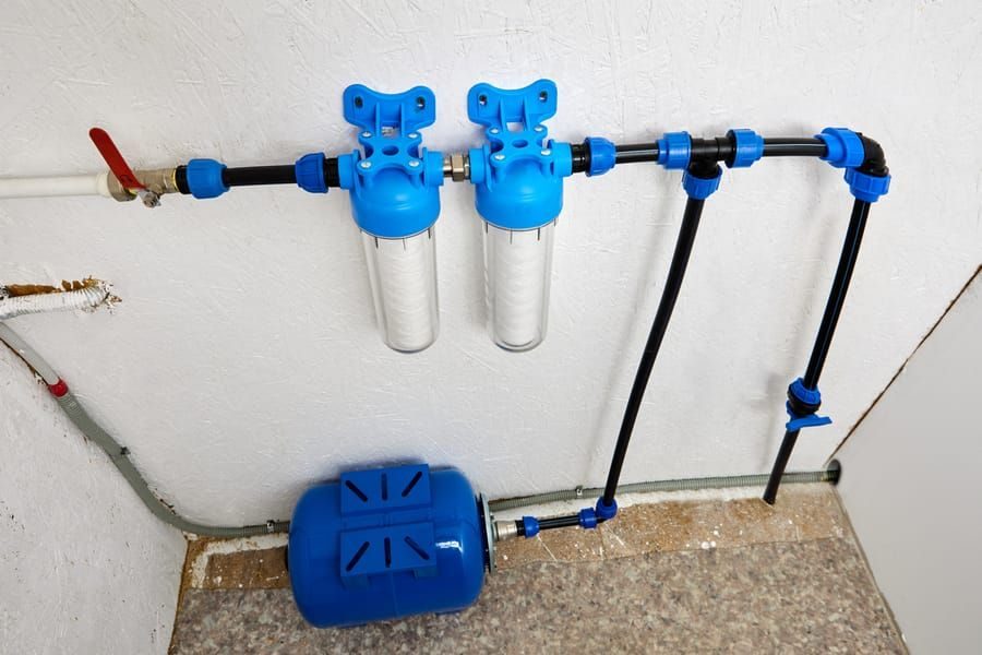 Blue home water filtration system with two inline filters and a wall-mounted pressure tank on the floor. Non-return pipework runs along a white wall.