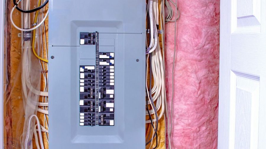 Electrical breaker panel mounted in an unfinished wall with many wires and pink insulation nearby.
