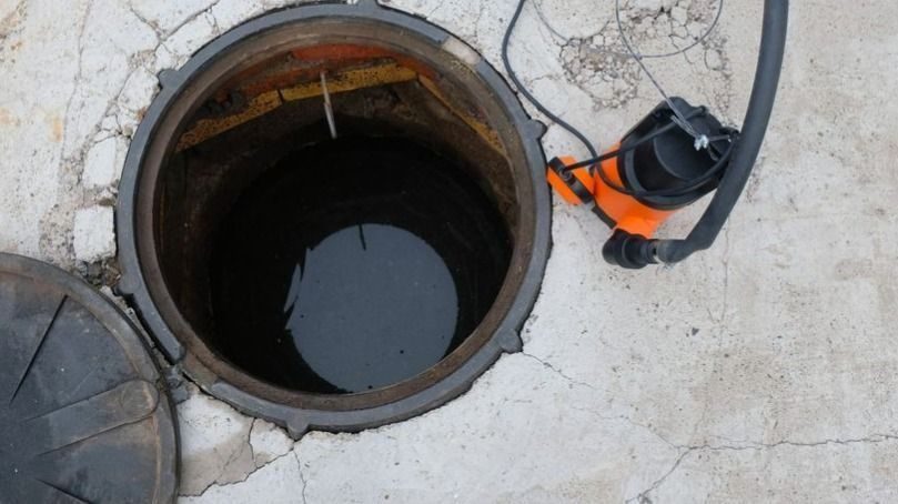 Open manhole exposing dark water on a cracked concrete surface; nearby manhole cover and an orange safety device with a black hose lie on the pavement.