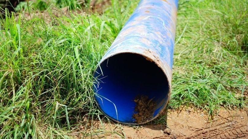 Blue plastic pipe lying on dirt and grass with its open end facing the camera, showing soil inside the tube.