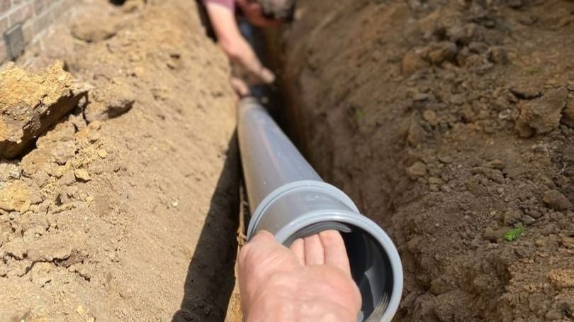 Workman inserts a gray PVC pipe into an excavated trench for installation or drainage.